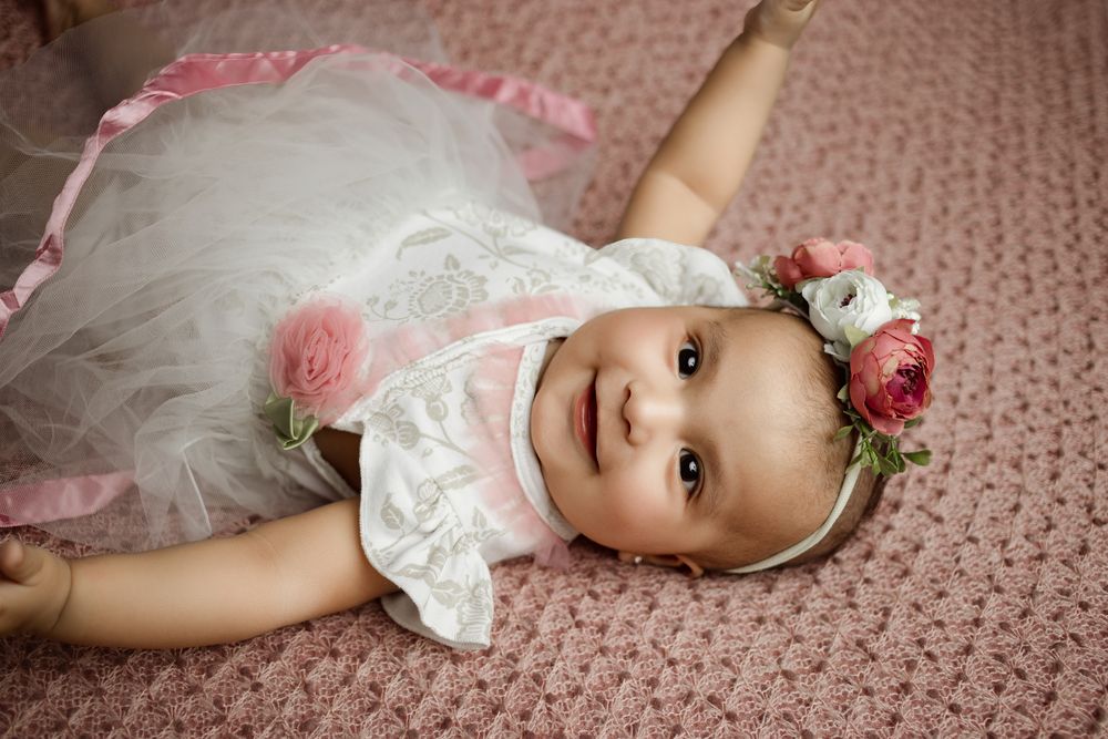 A little one wearing a white lace dress and floral headband lies on a pink textured carpet.