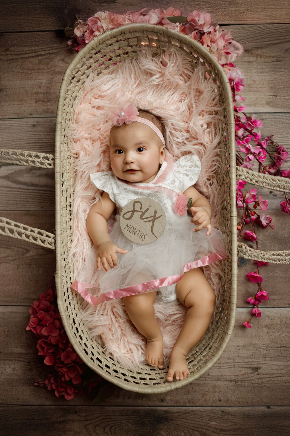 Wicker bassinet decorated with pink roses and flowers on wooden floor contains soft pink blanket.