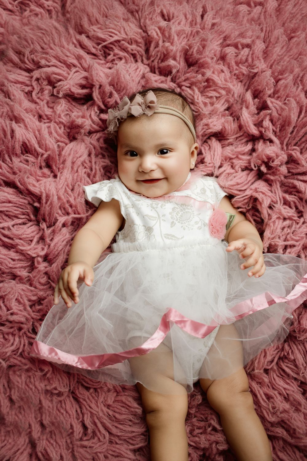 Baby wearing white tulle dress and headband lies on pink fluffy rug, smiling brightly.