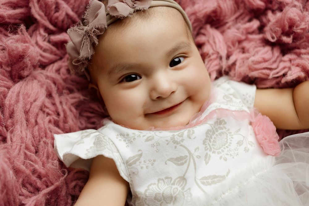 A baby wearing a white lace dress and bow headband smiles while lying on pink textured fabric.