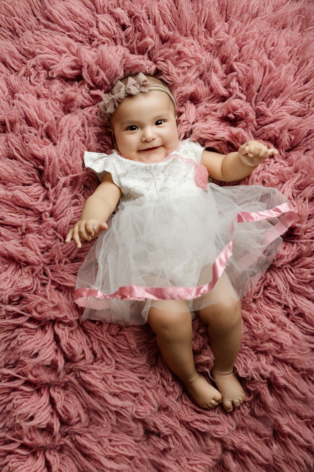 Baby wearing white lace dress smiling while lying on pink fluffy rug background.