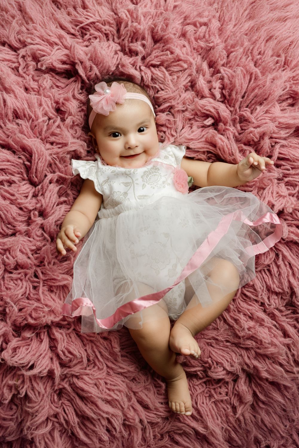 Baby in white tulle dress lying on pink fluffy rug during professional photo shoot.