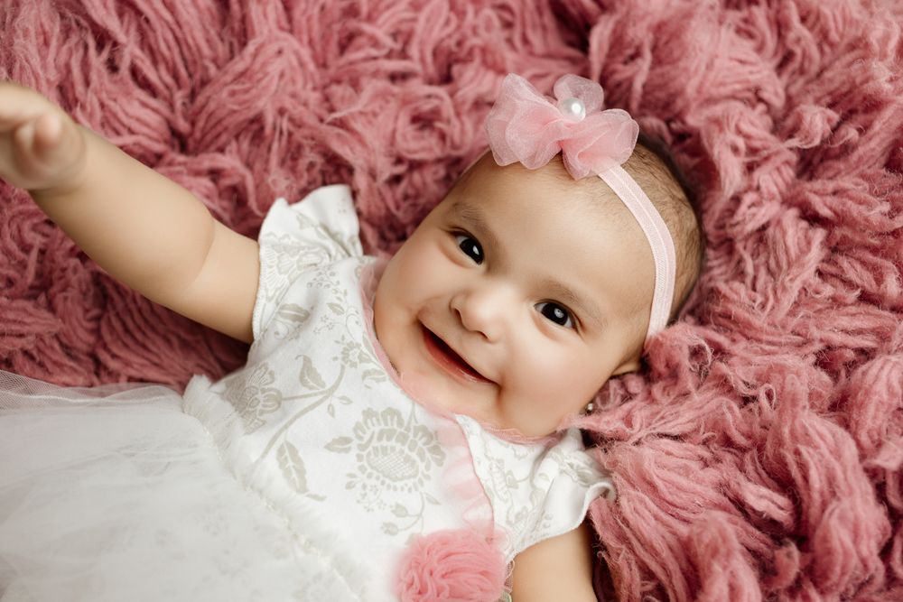 Baby wearing white lacy dress and pink bow lying on plush pink rug with big smile.
