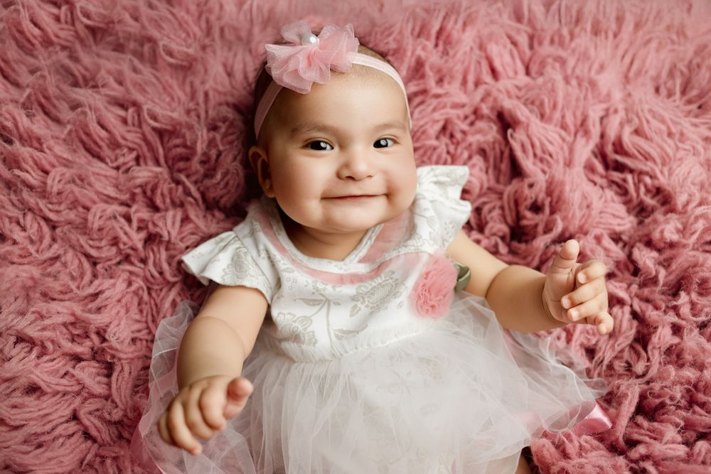 Baby in white dress smiles while lying on fluffy pink blanket with matching pink bow accessory.