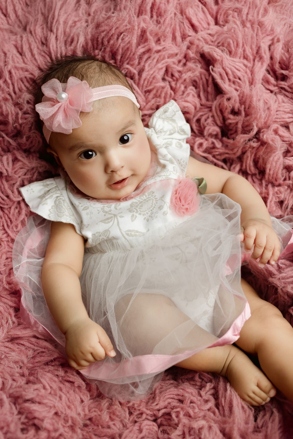 Baby wearing white dress with pink bow headband lying on pink fluffy rug during professional photo session.