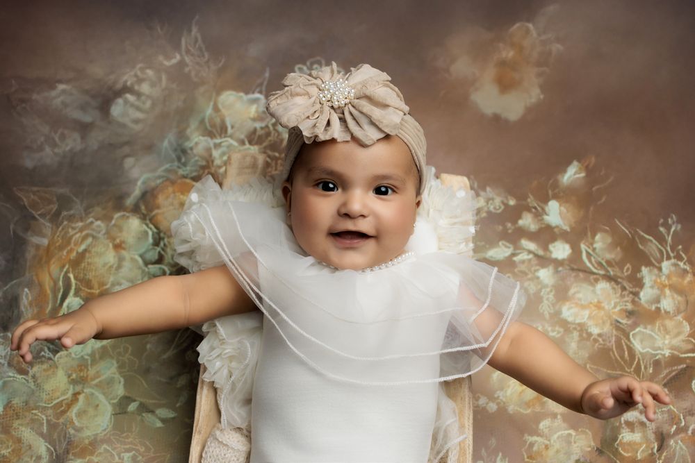 Ethereal portrait of a baby in white ruffled dress and floral headpiece against dreamy pastel background.