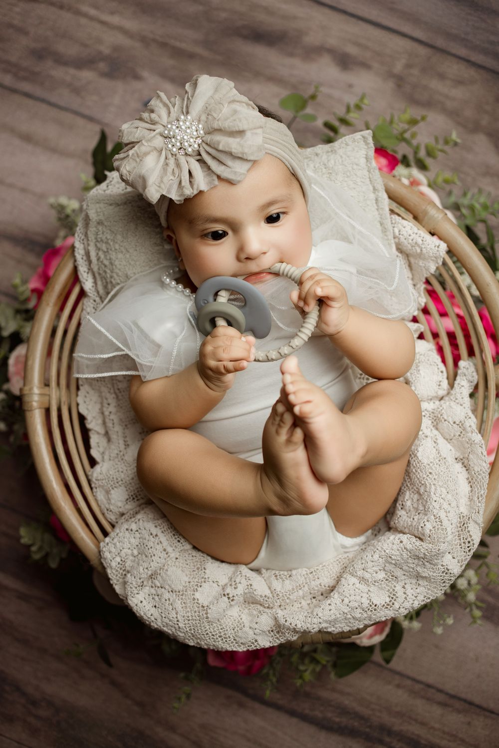 Baby in white dress with floral headpiece lying in woven basket with soft white bedding and pink floral accents.