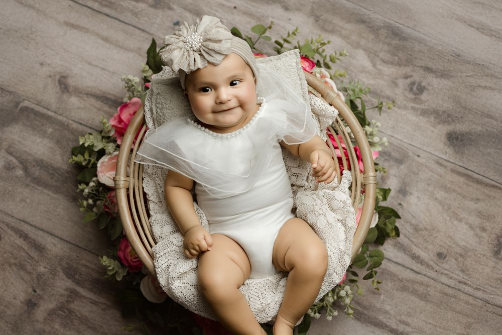 Baby in white outfit smiling while posed in rustic basket with floral decorations.
