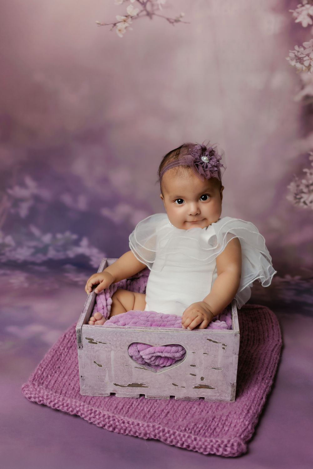 Baby in white dress sitting in vintage wooden box on pink knitted blanket with magical purple backdrop.