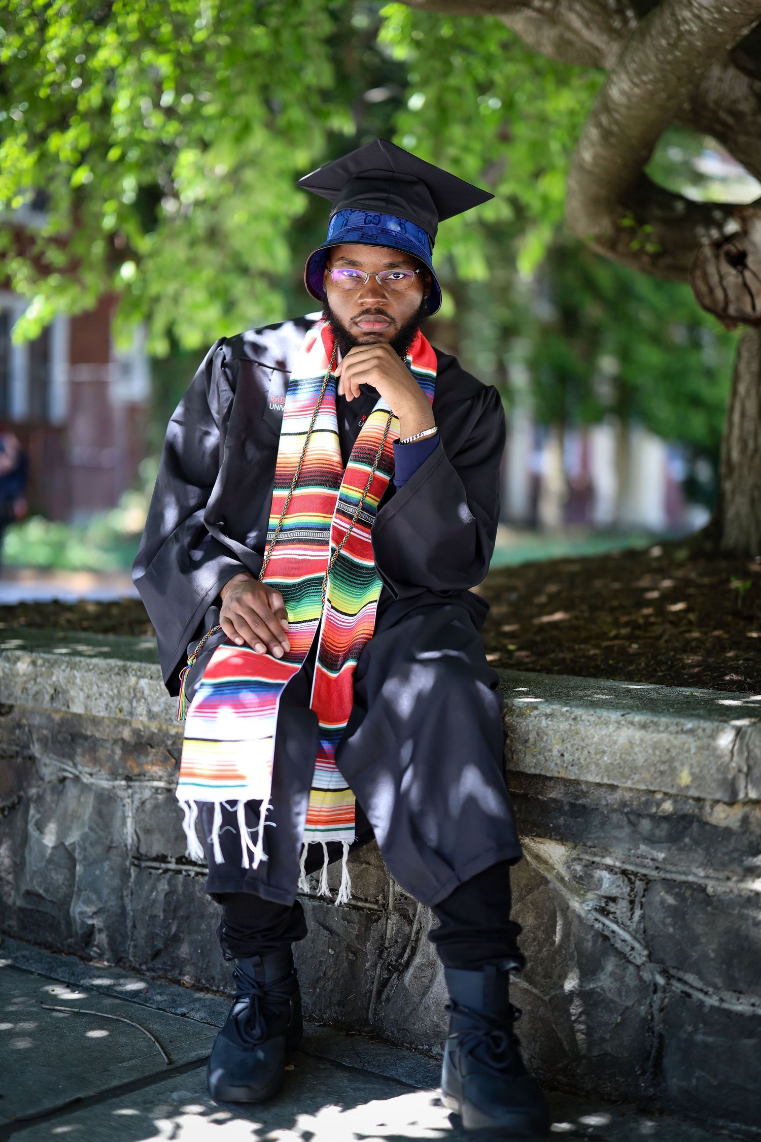 Graduate in cap and gown with colorful kente stole sitting thoughtfully in dappled sunlight.
