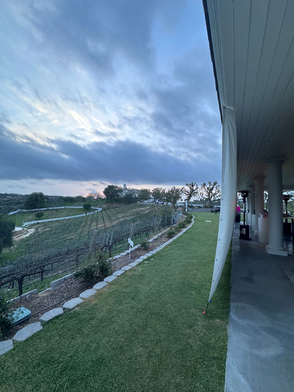 Scenic vineyard view from a covered patio with dramatic cloudy sky at sunset and well-maintained landscaping.