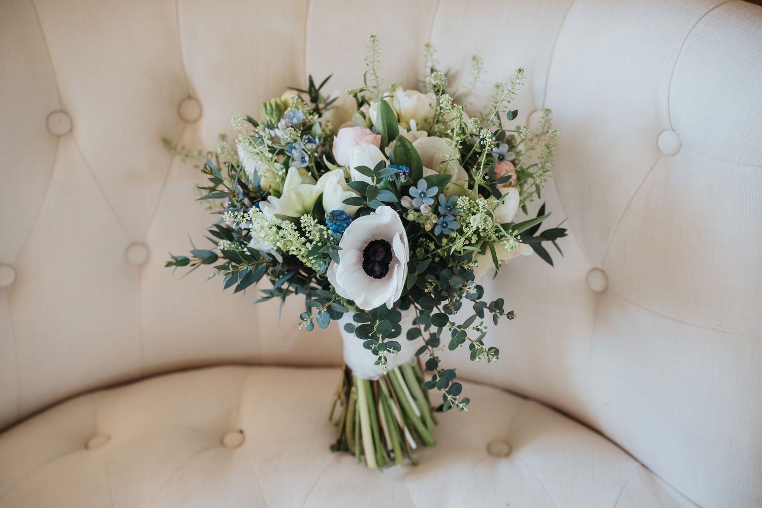 A bridal bouquet of white anemones, blue flowers, and eucalyptus rests on a cream tufted chair.