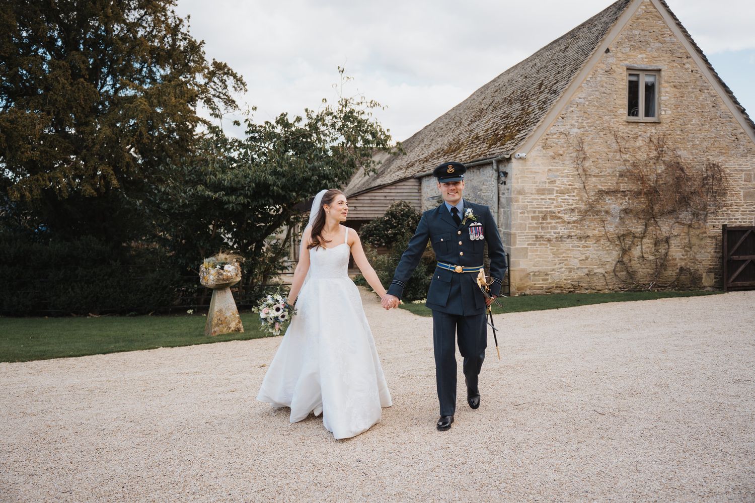 A bride in a white gown walks hand in hand with a groom in military uniform outdoors.
