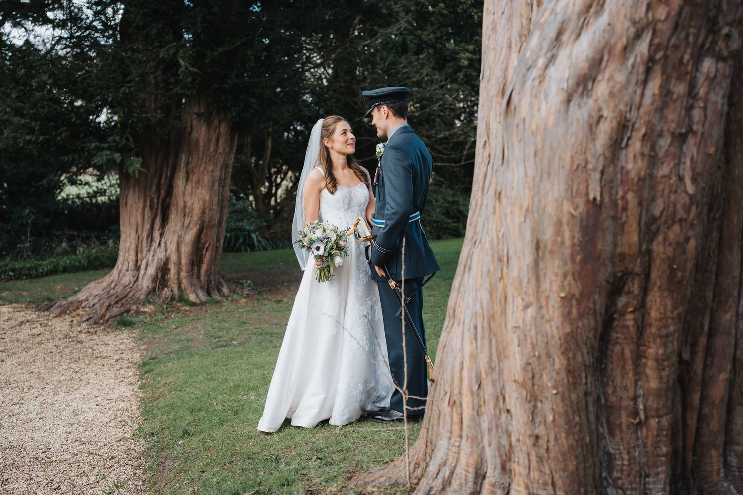 A bride and groom share a tender glance while standing between tall ancient trees in a garden.