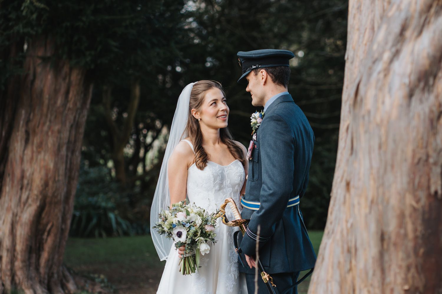 Bride and groom share a romantic moment among tall trees, she holds a white bouquet as he wears a military dress uniform.