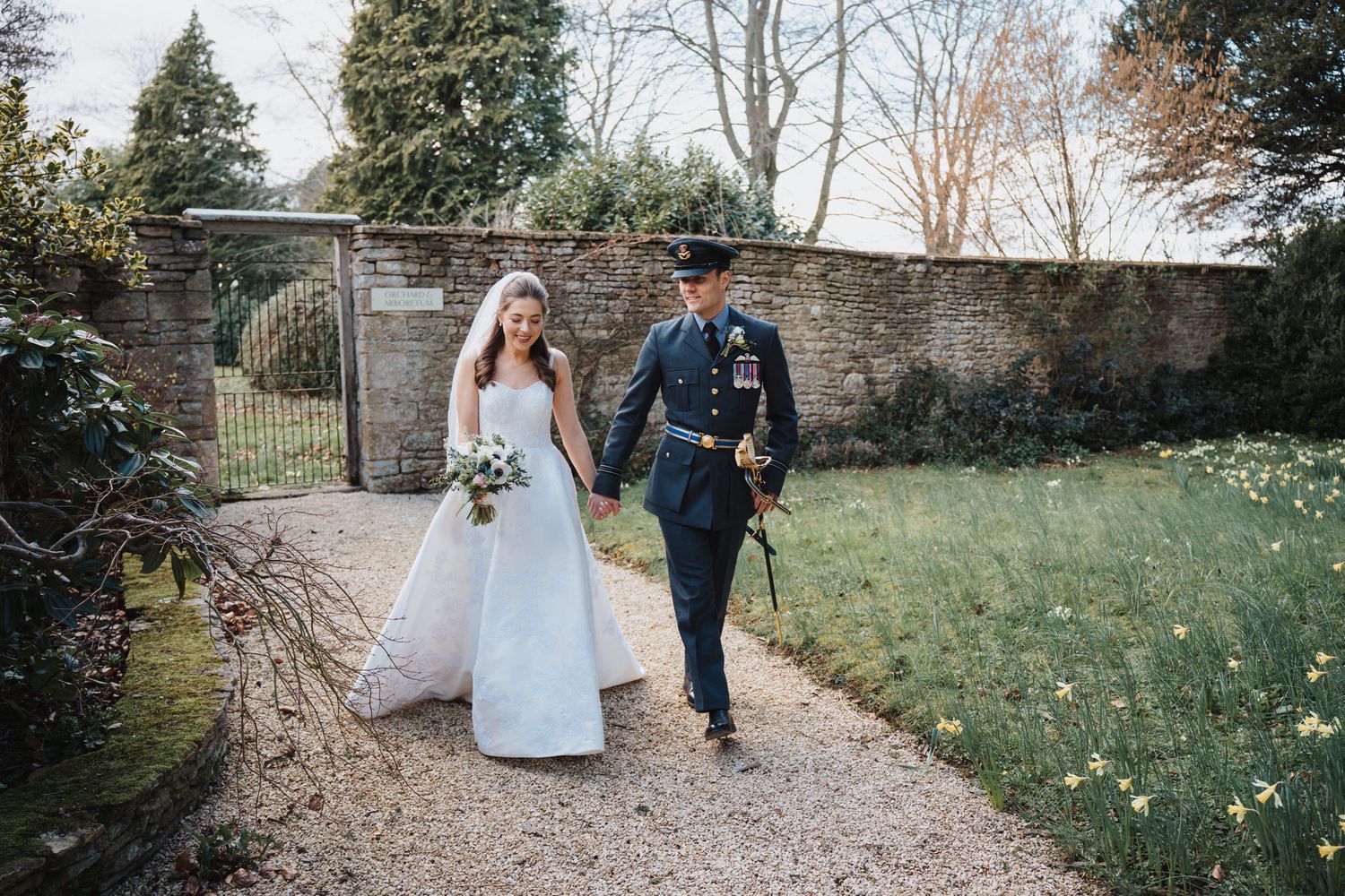 Newlywed couple walks hand in hand along a garden path, bride in white gown and groom in military uniform at a countryside venue.