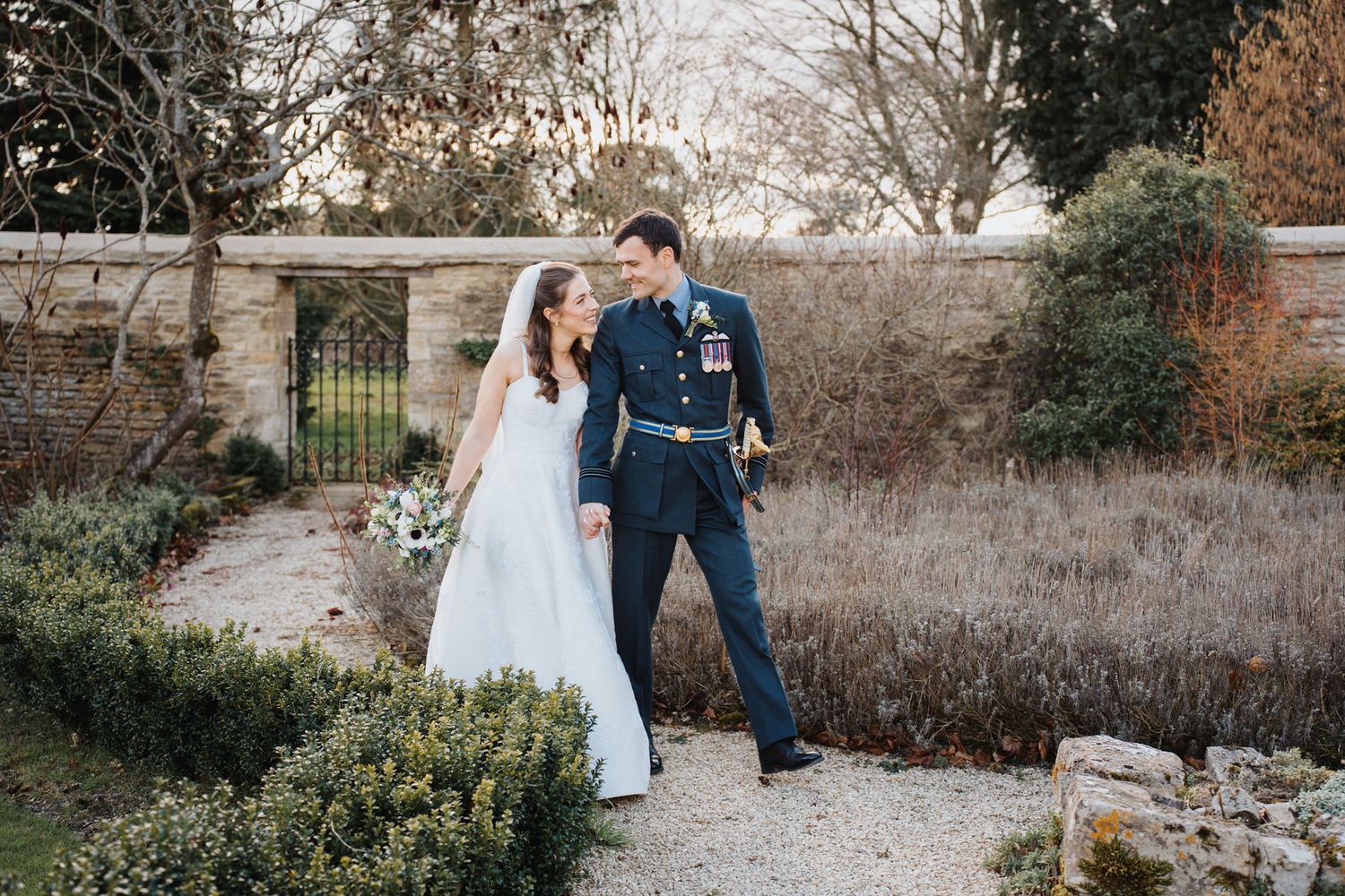 Smiling bride and groom in military uniform stroll through a walled garden with gravel paths and winter foliage.