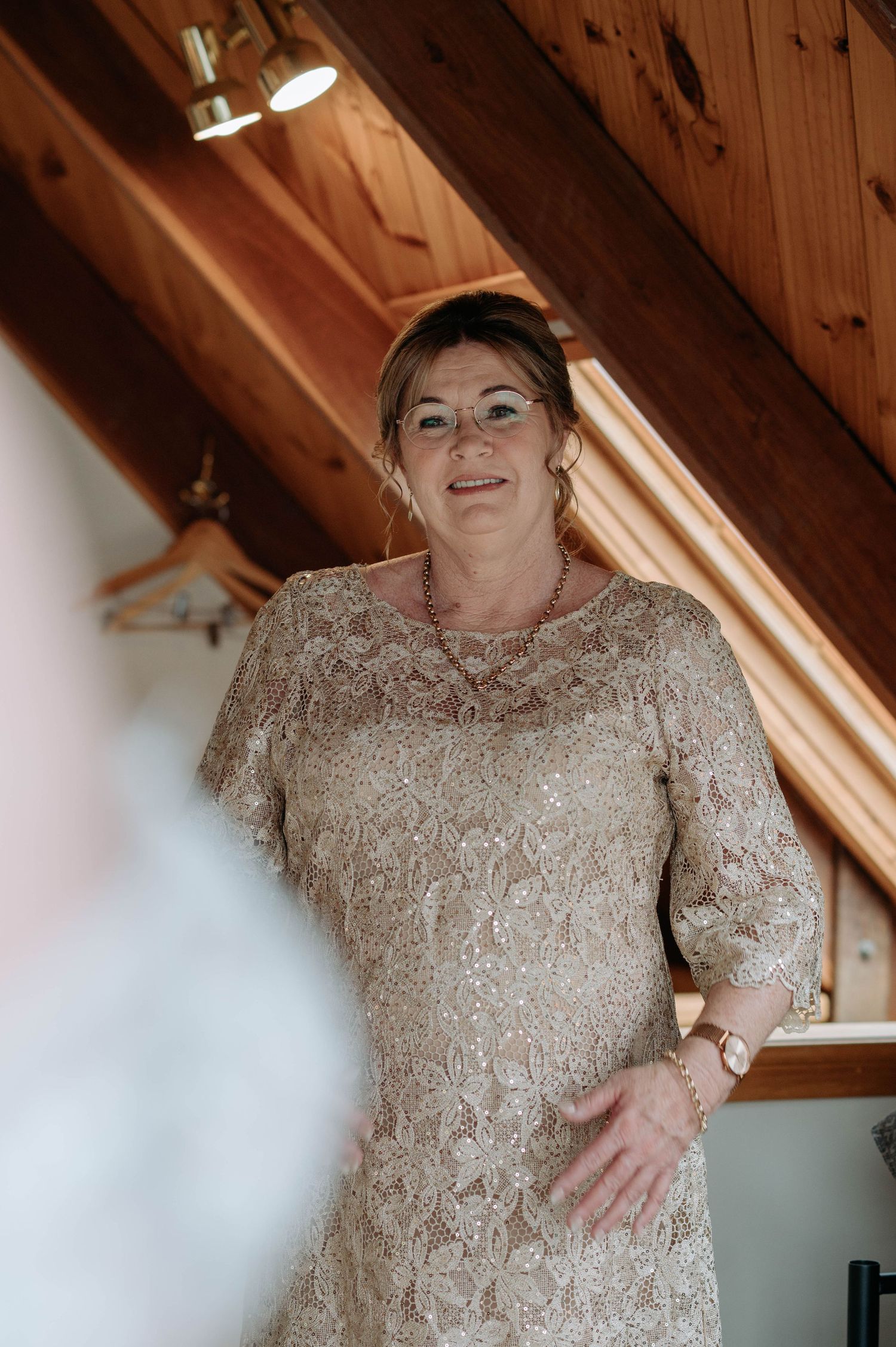 A woman wearing a sparkly beige lace dress stands in an attic room with wooden ceiling beams.