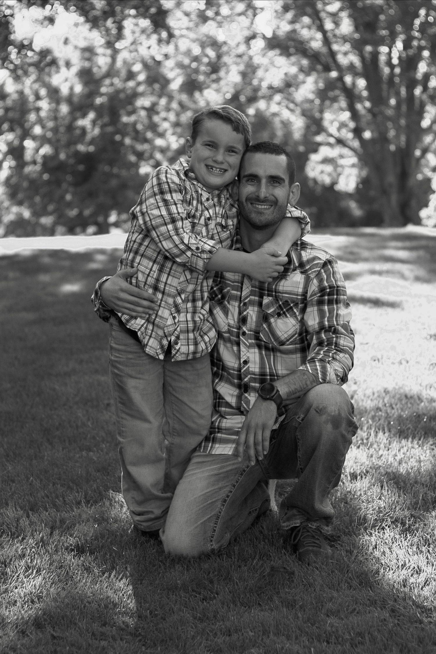 Black and white portrait of a father and son in matching plaid shirts outdoors.