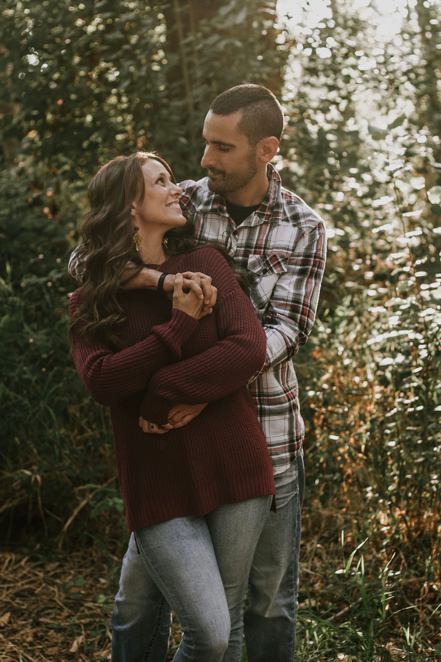 Couple embracing and smiling in autumn forest, woman in burgundy sweater, man in plaid shirt.