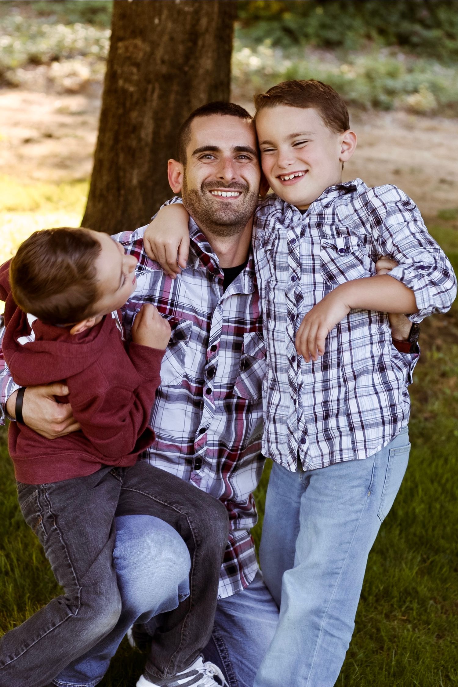Smiling father in plaid shirt hugging two young boys outdoors near a tree.