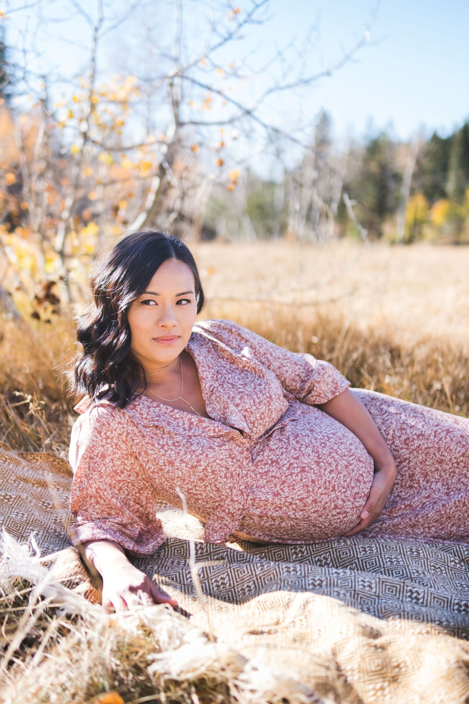 Maternity photo in fall field shows expecting mother wearing floral pink dress reclining on blanket among autumn grasses.
