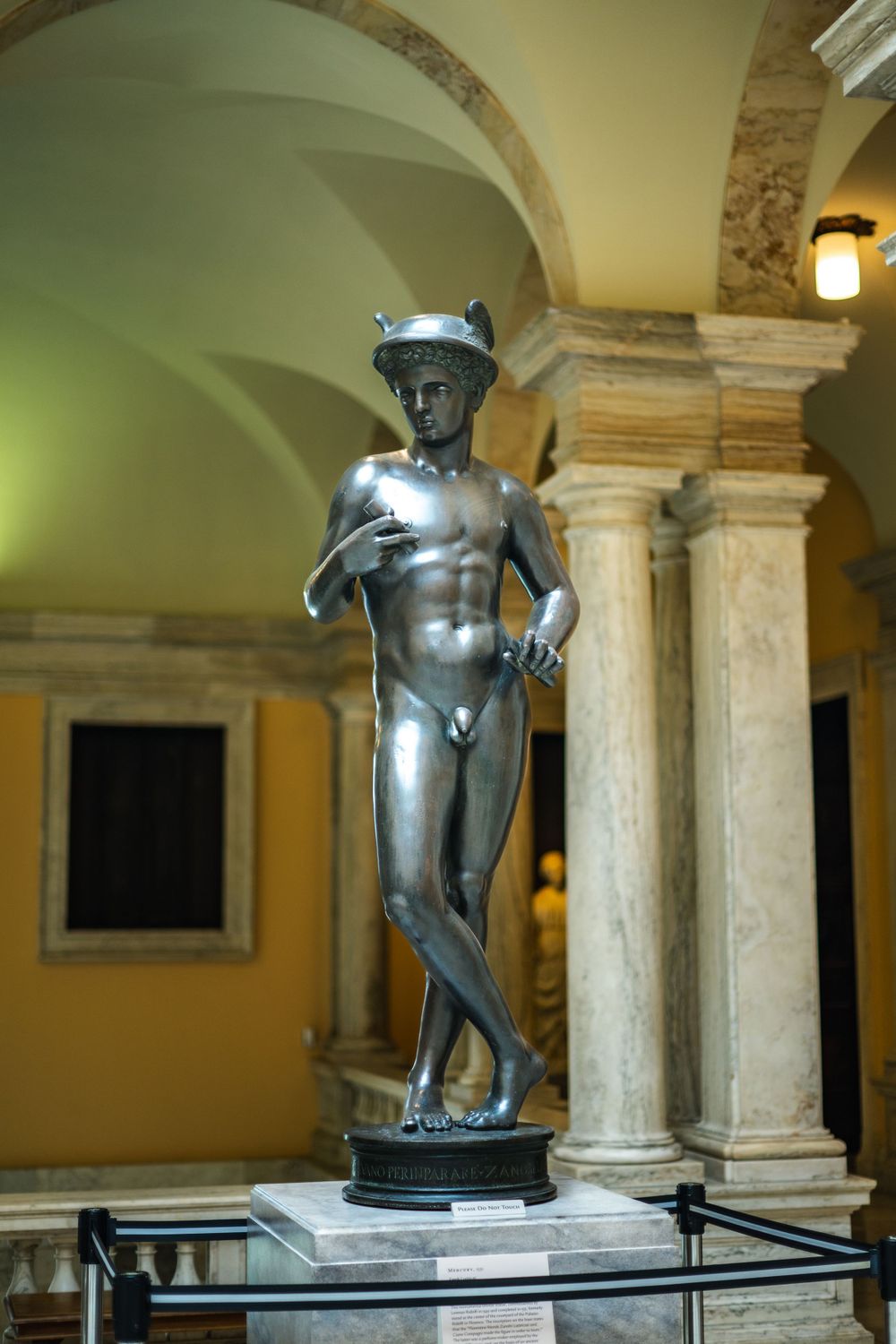 Bronze statue of Mercury displayed in a classical interior with yellow walls and stone columns illuminated by warm lighting.
