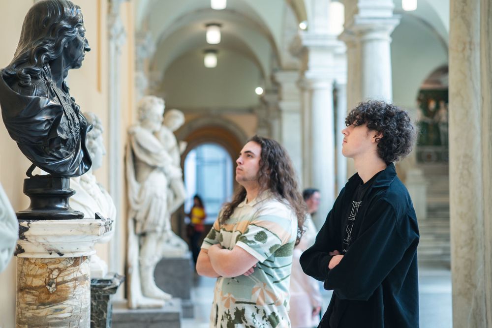 Two visitors admire classical art and sculptures displayed in an ornate museum gallery with arched columns and marble busts.