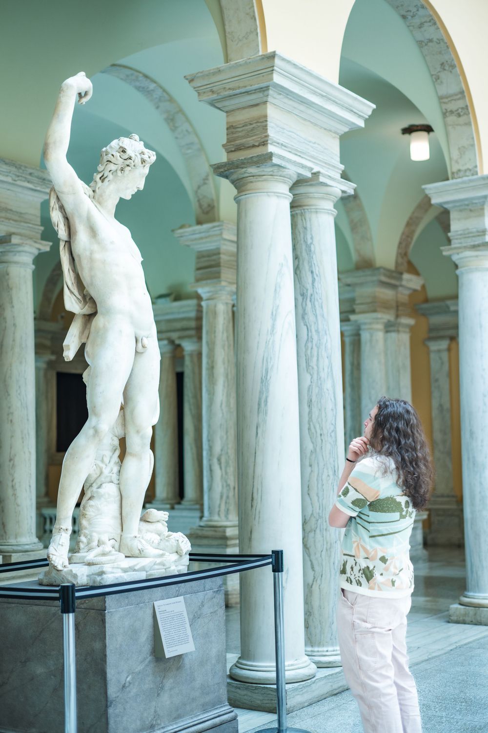 Museum visitor observes a classical marble statue in an elegant colonnade with turquoise walls and white columns.