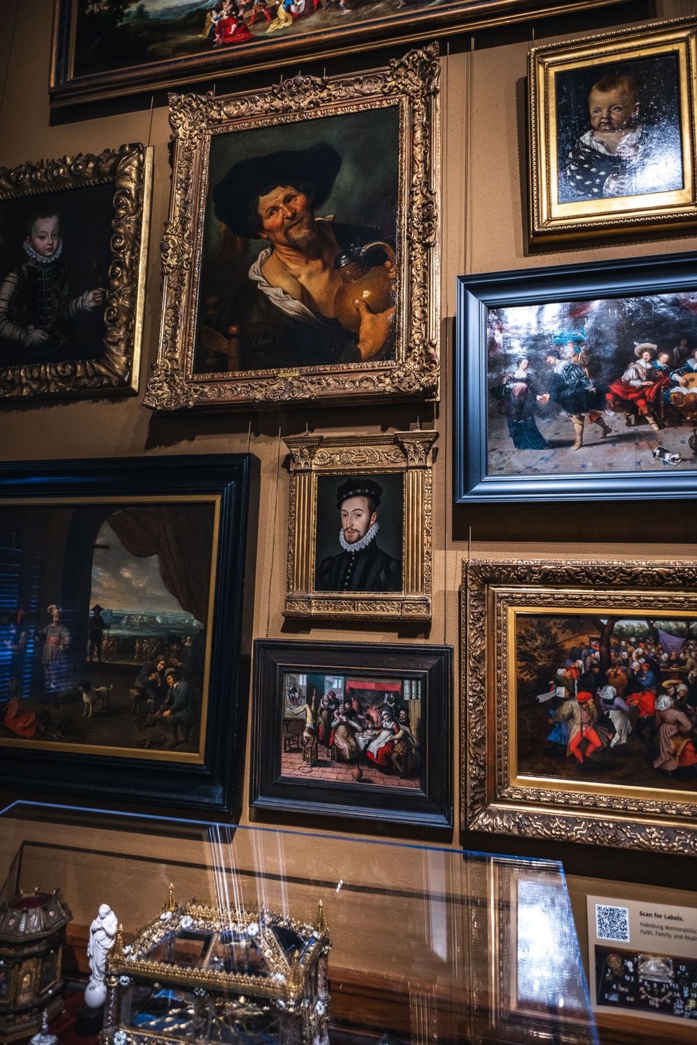 A wall display of ornate gilded frames containing classical paintings in a traditional gallery or museum setting.