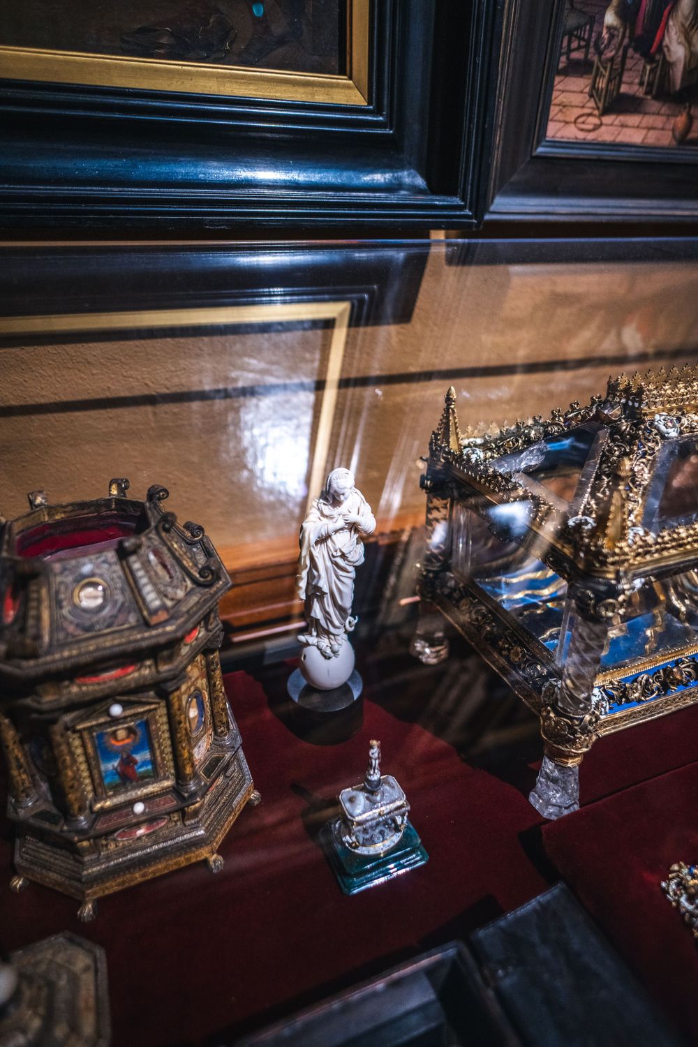 Ornate antique religious relics and decorative objects displayed on red velvet shelf including carved statues and gilded boxes.