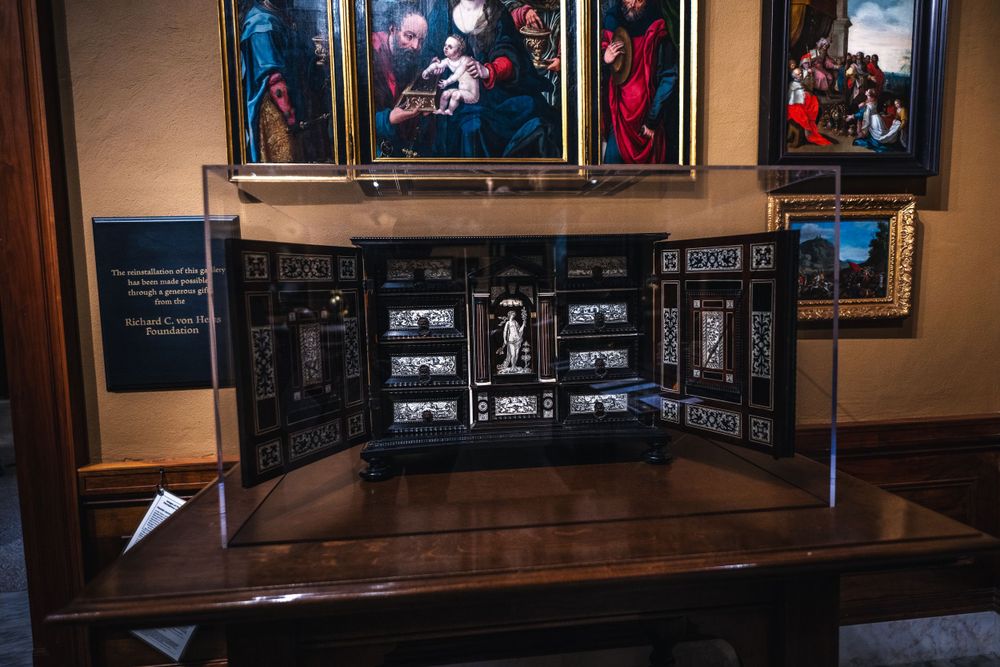 A wooden display case with historic photographs and documents beneath framed classical paintings in a museum gallery.