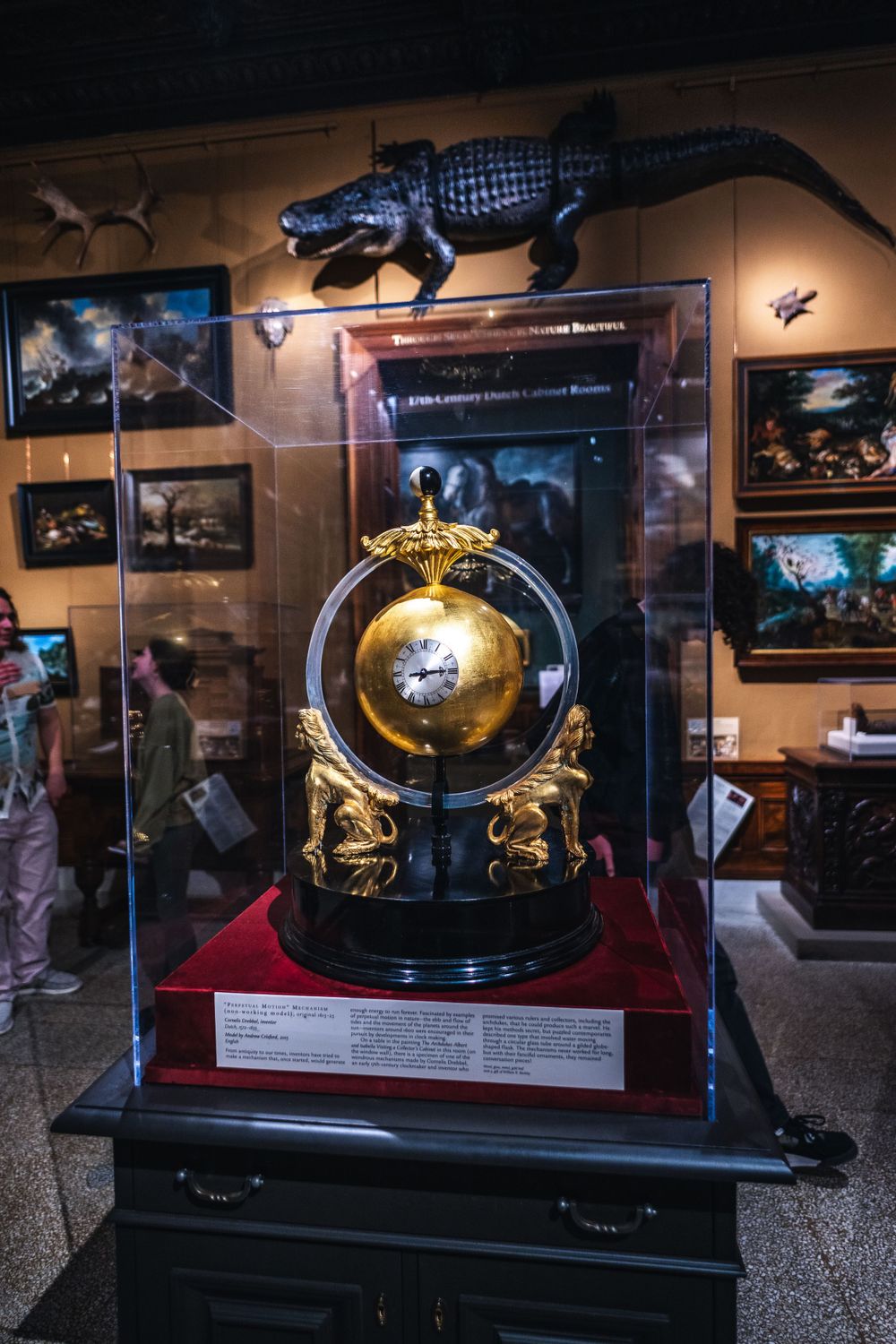 Ornate golden globe display in glass case at museum with dramatic lighting and dinosaur exhibits on the walls.