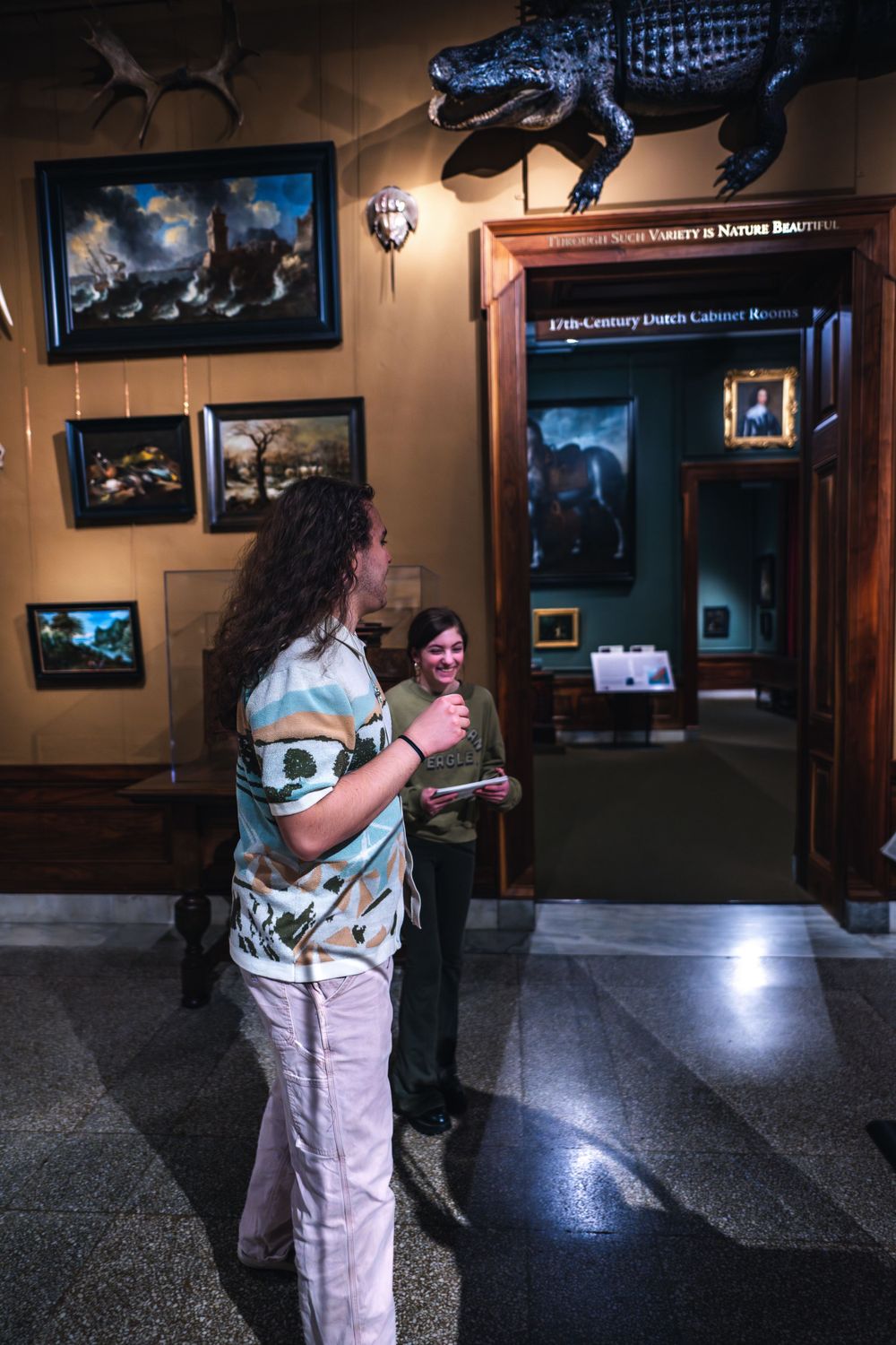 Two people stand chatting in a museum hallway with framed artwork and an alligator sculpture mounted on the wall.