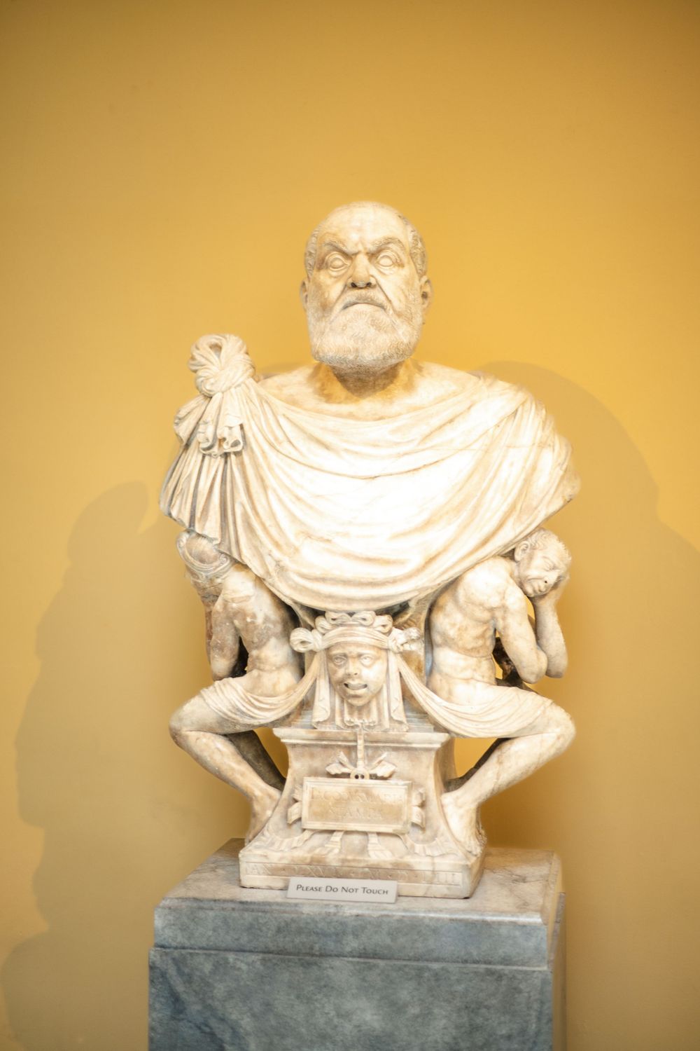 Ancient marble bust sculpture with draped toga and ornate pedestal base against beige wall.