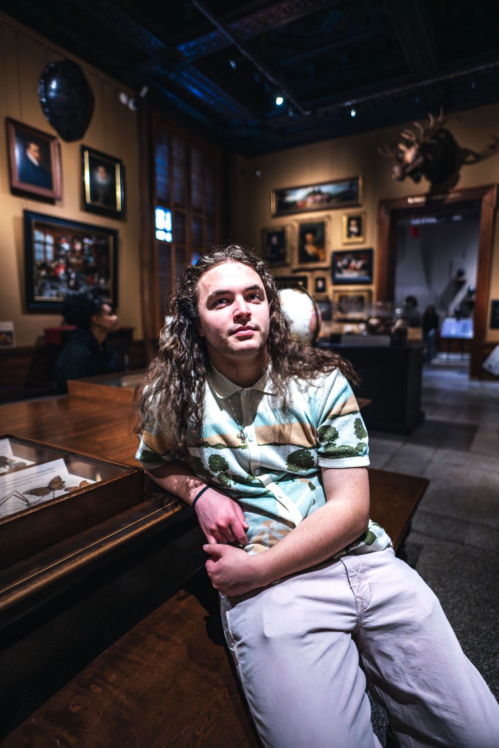 Person with long wavy hair wearing a striped polo shirt sits casually in an art gallery with framed pictures on walls.