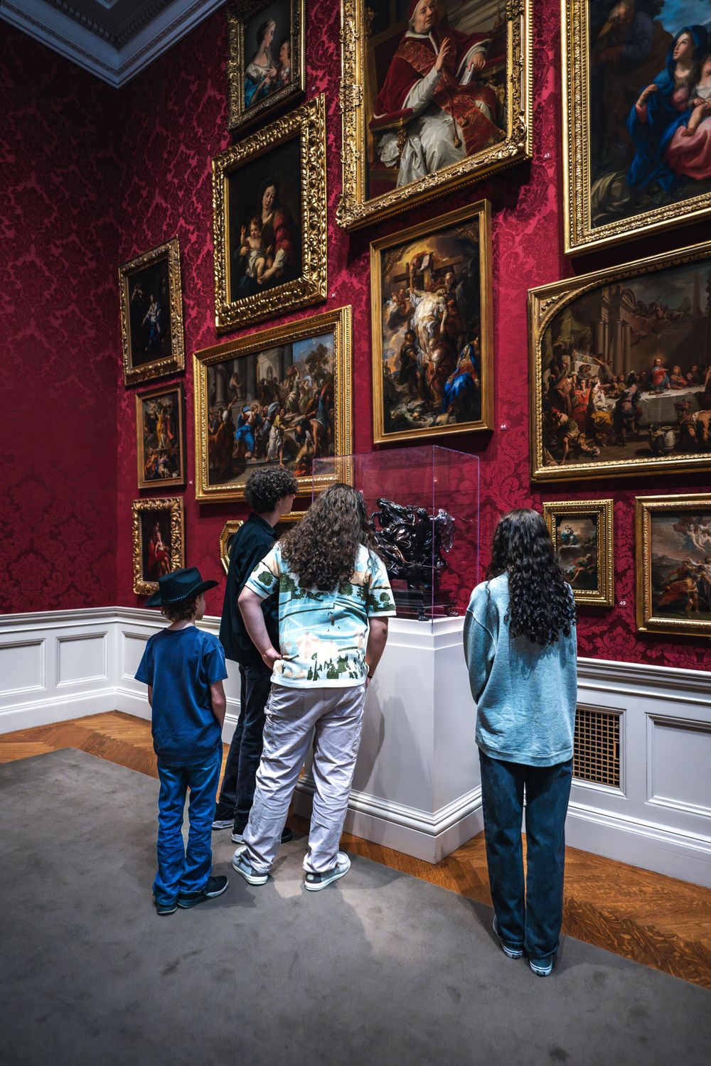 Museum visitors admiring classical paintings on red damask walls in an ornate gallery interior.