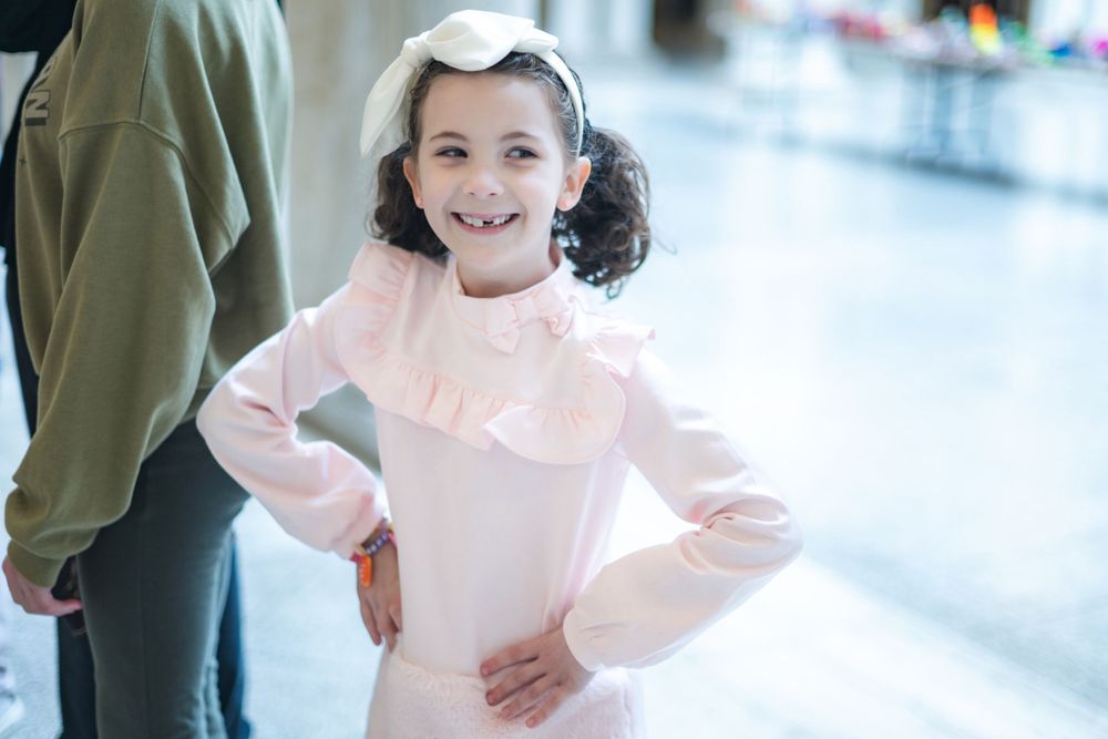 A young child wearing a light pink ruffled dress poses with hands on hips in an indoor setting.