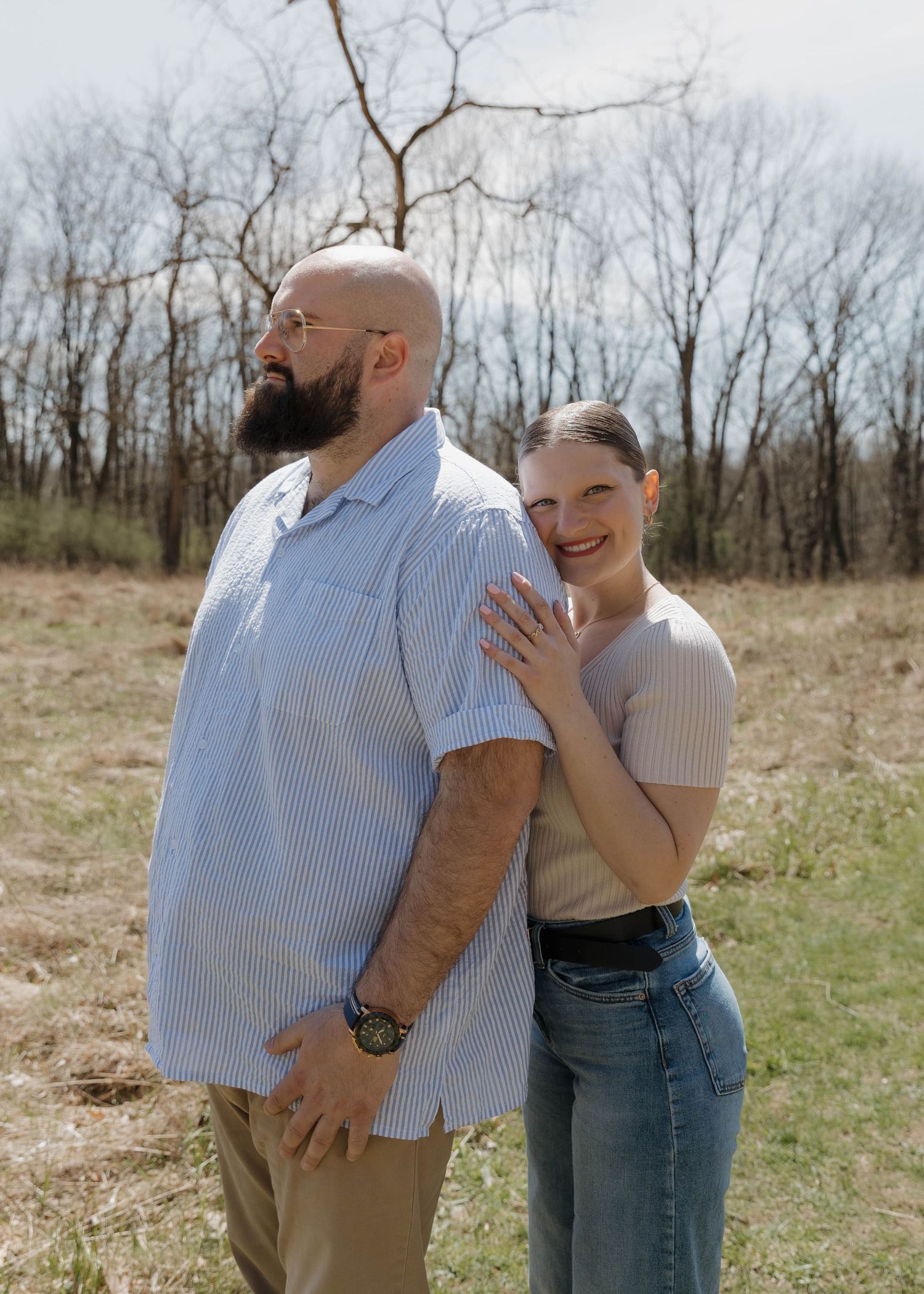 A couple poses outdoors in a field with bare trees in the background on a cloudy day.