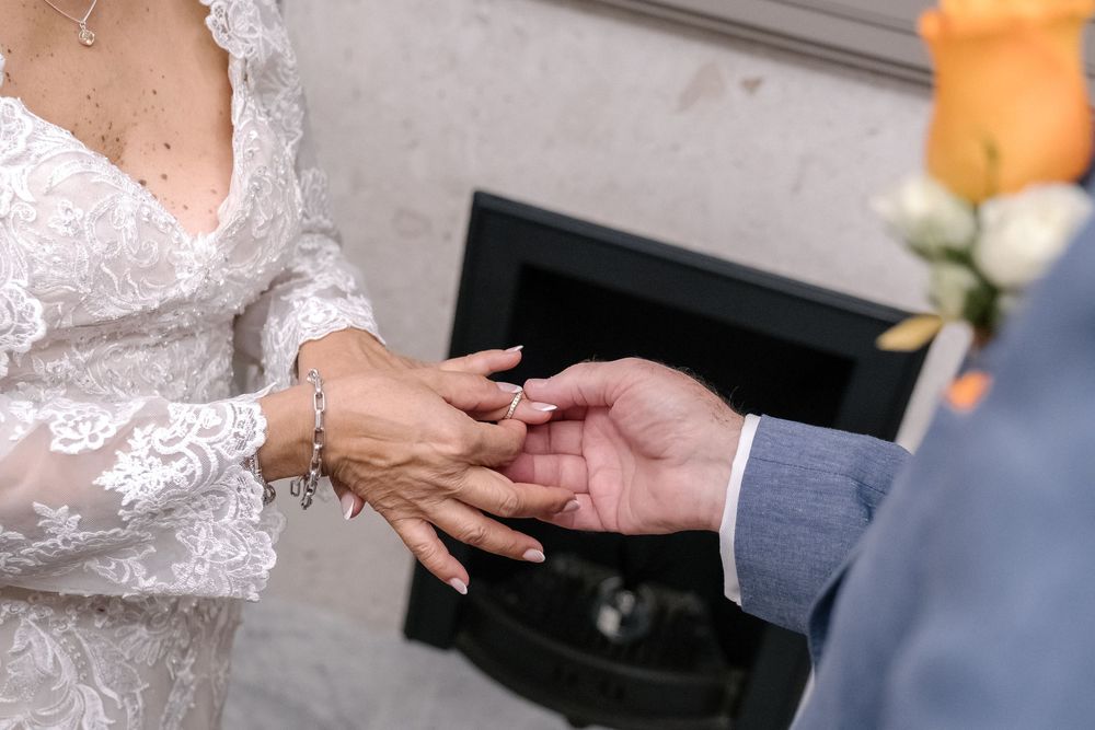 Close-up of hands during ring exchange at wedding ceremony showing delicate lace sleeve and gray suit.