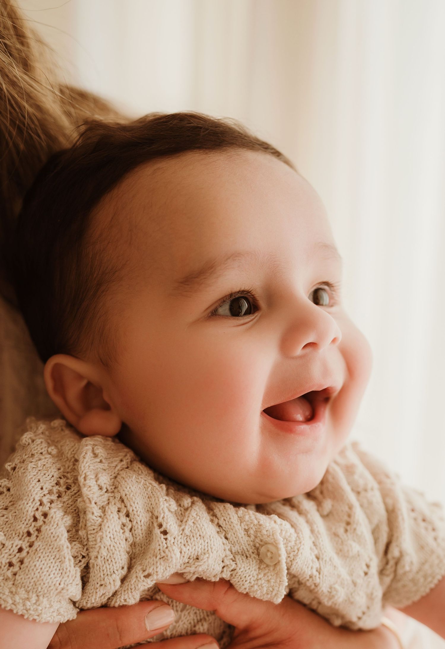Sweet baby in cream knit sweater sits on textured blanket near pampas grass in warm neutral toned nursery setting.