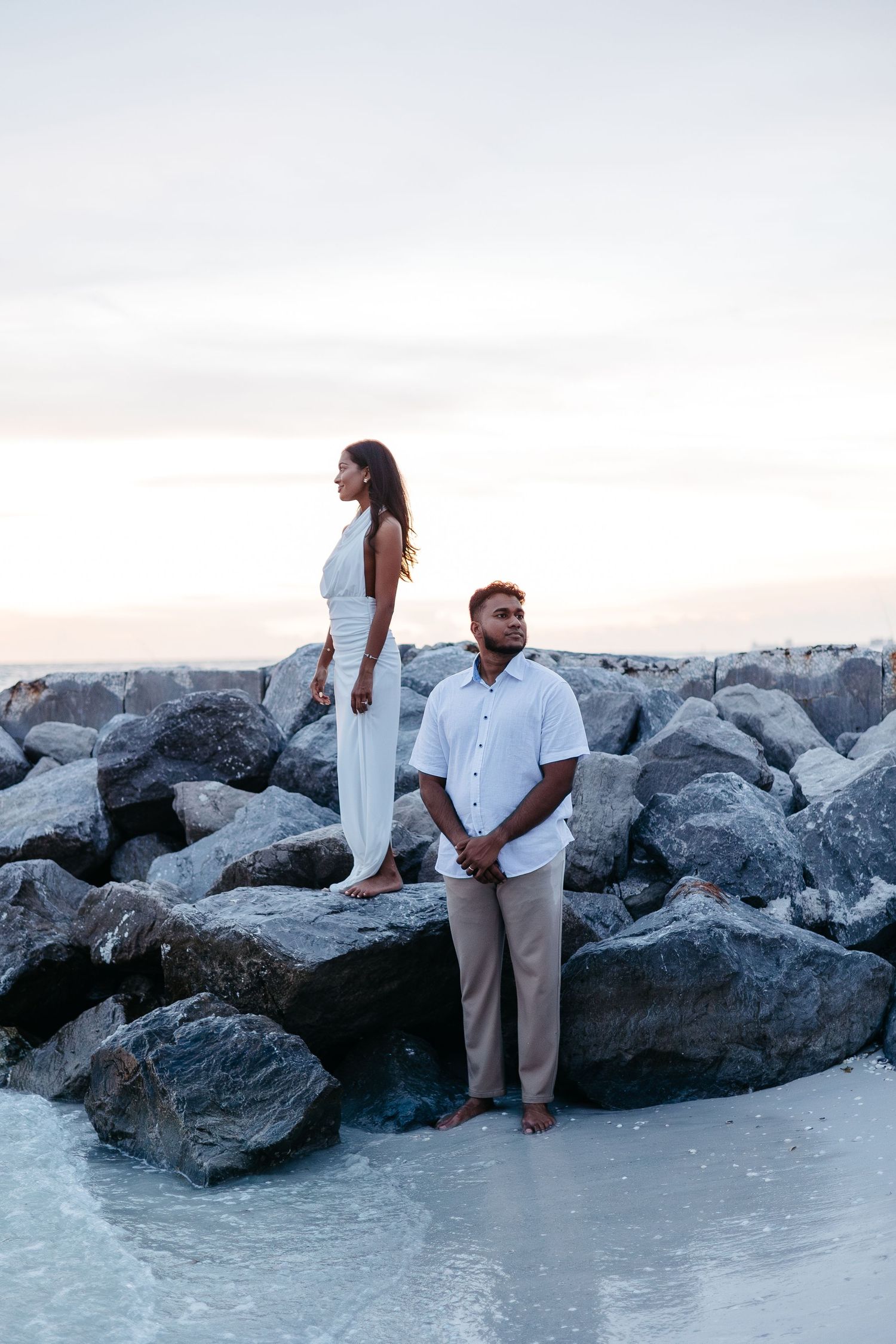 Couple posing on rocky beach during sunset in casual white outfits.