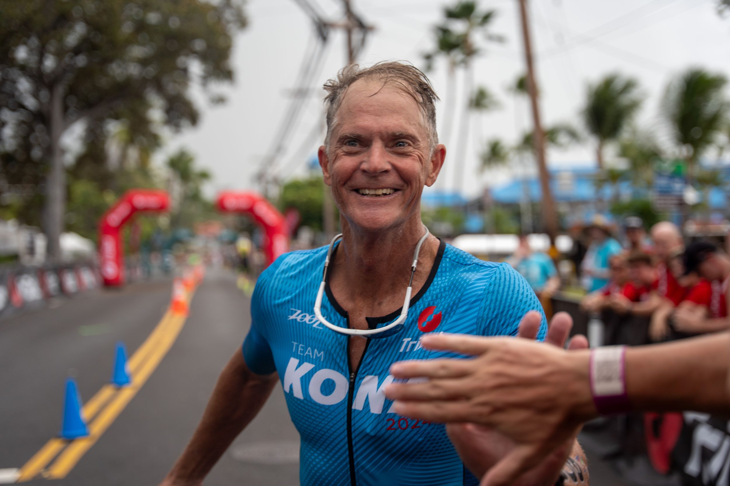 Athlete in light blue KONA jersey smiles during outdoor sporting event with palm trees and crowd barriers visible.