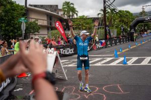 Athlete in blue triathlon gear celebrates with raised arms at finish line during race event on city street.