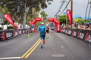 Runner approaching finish line with arms raised between red banners at outdoor race event on city street.