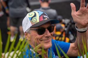 Participant in athletic wear and sunglasses waves while wearing a colorful patterned cap at outdoor sporting event.