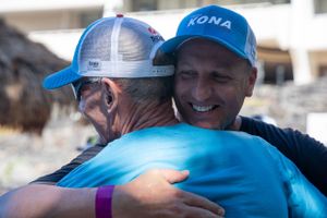 Two people wearing blue KONA caps share a warm embrace and smile during an outdoor event.