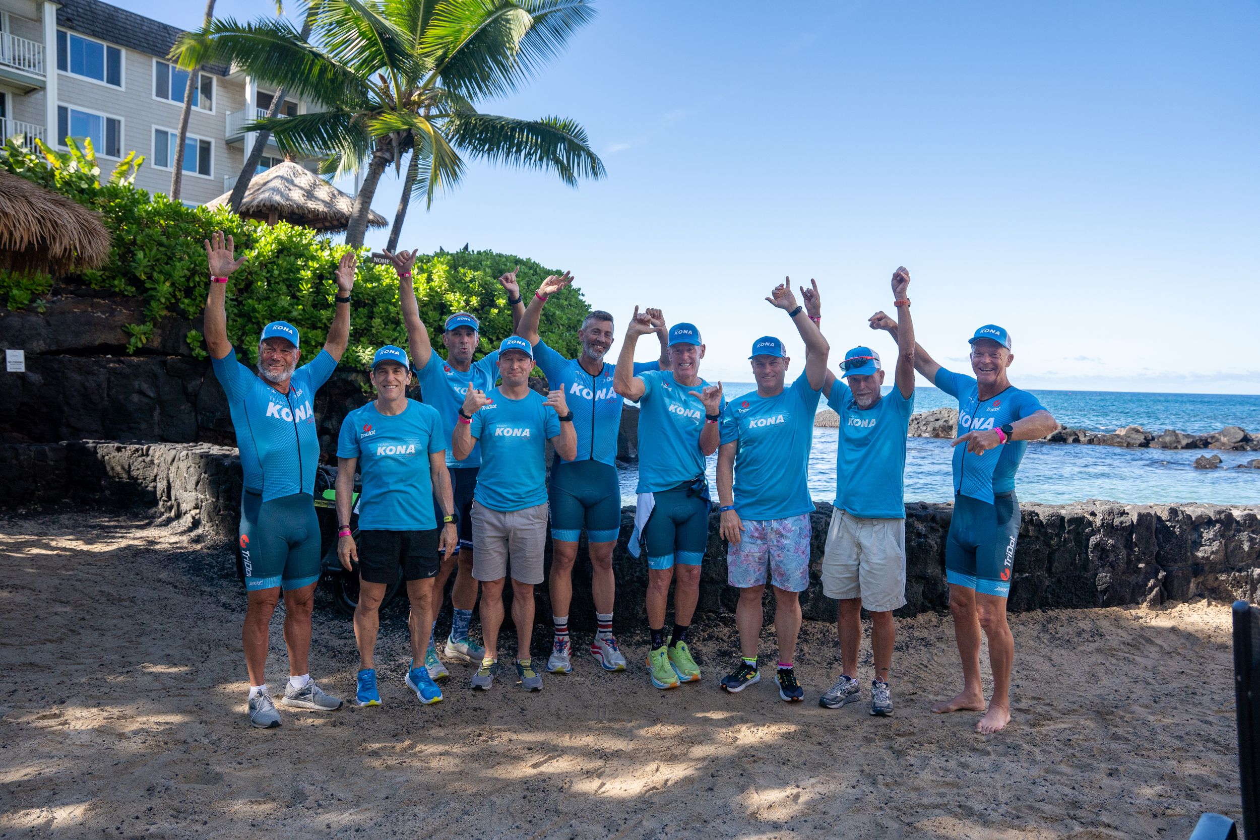 A group in matching blue t-shirts stands together on a tropical beach with palm trees and ocean views in Hawaii.