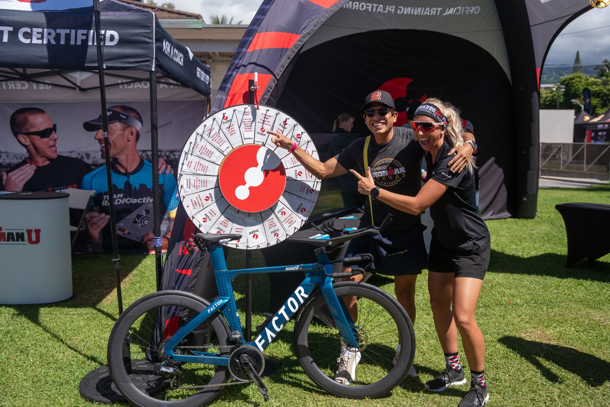 People pose with a blue mountain bike and spinning prize wheel at an outdoor event tent display.