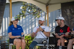 Three people in casual attire and baseball caps sit together having a conversation under a covered outdoor area.